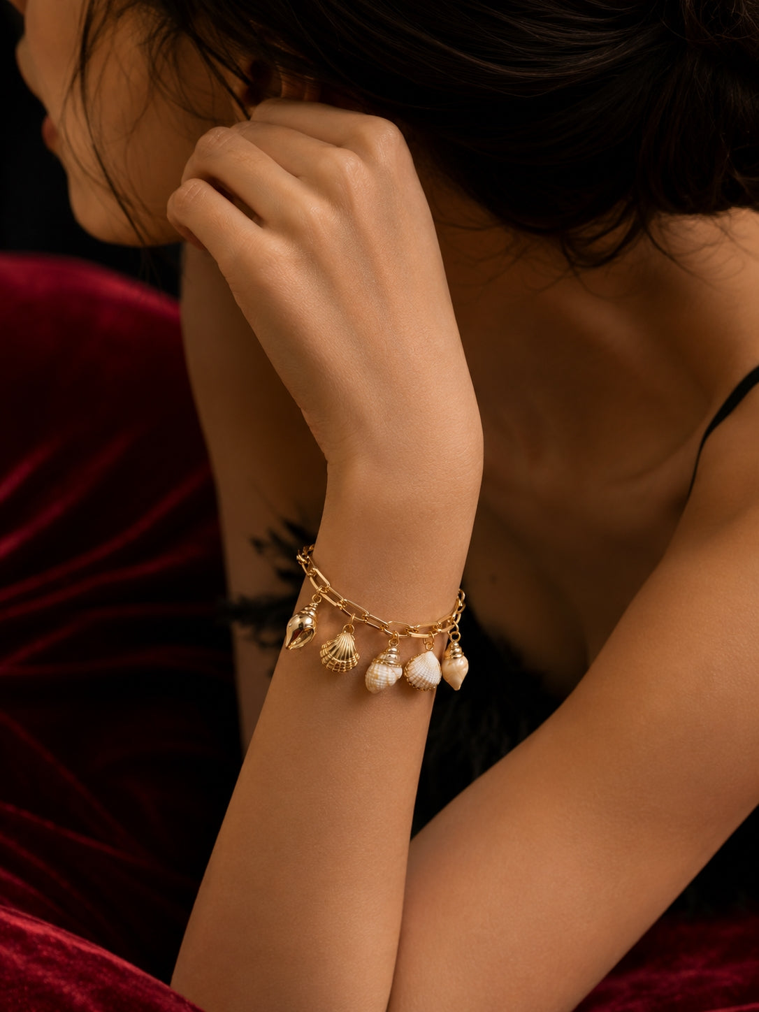 Model wearing gold seashell charm bracelet on wrist against red velvet